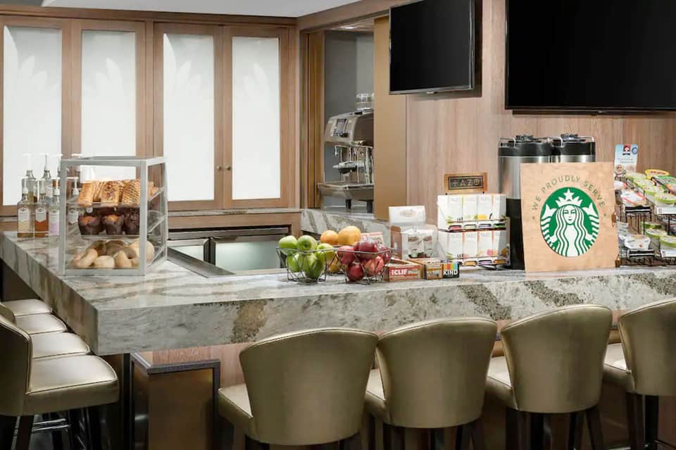 Hotel breakfast area with marble countertop, Starbucks signage, pastries, fresh fruit, and wood-paneled walls