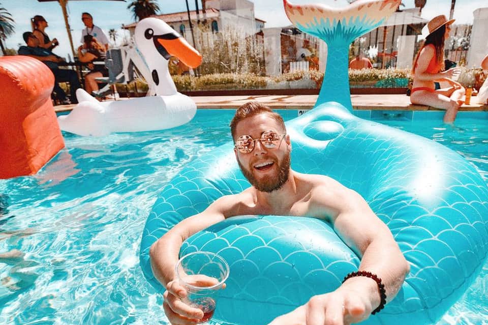 Man on turquoise mermaid float in resort pool with flamingo float and colorful buildings in background