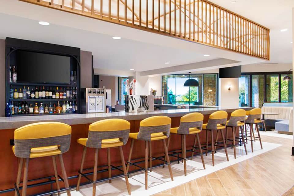 Modern hotel bar with yellow upholstered stools, wooden ceiling detail, and full liquor display