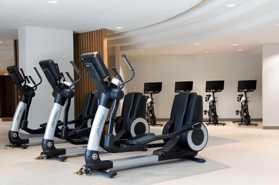 Modern fitness center with elliptical machines and stationary bikes with monitors in bright white space