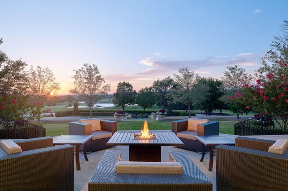 Fire pit seating area with woven chairs and pink flowers overlooking a sunset landscape