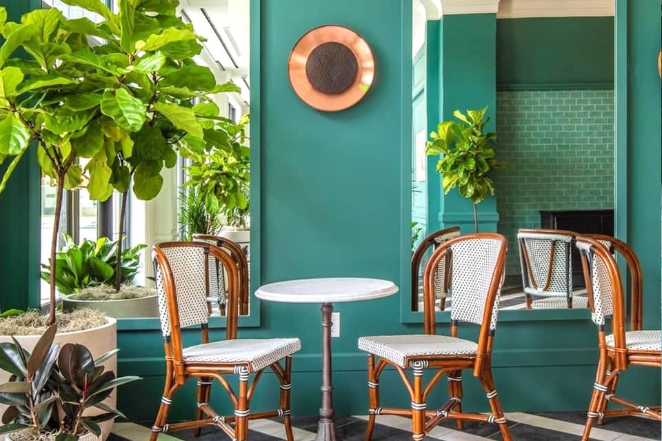 Turquoise outdoor café with rattan chairs, small round table, potted plants, and coral-colored speaker on wall