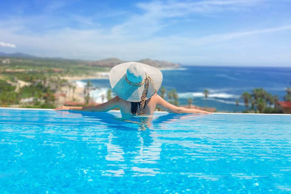Woman in wide-brimmed hat relaxing in infinity pool overlooking tropical coastal landscape
