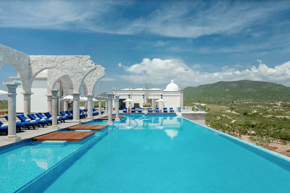 Luxury resort infinity pool with white columns and arches, blue loungers, overlooking desert landscape and mountains