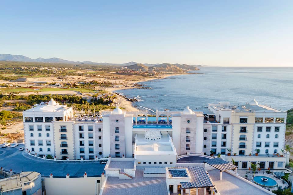 Aerial view of white resort buildings with pools overlooking Mediterranean coastline and mountains