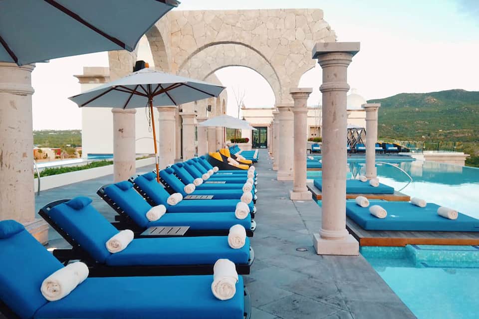 Row of blue lounge chairs under white umbrellas on stone patio with classical columns and turquoise pool