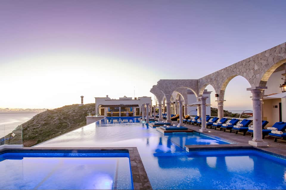 Luxury resort infinity pool at dusk with arched colonnade, loungers, and coastal hillside views