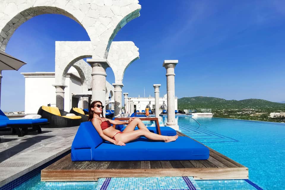 Woman lounging on blue daybed by infinity pool with white columns and ocean view
