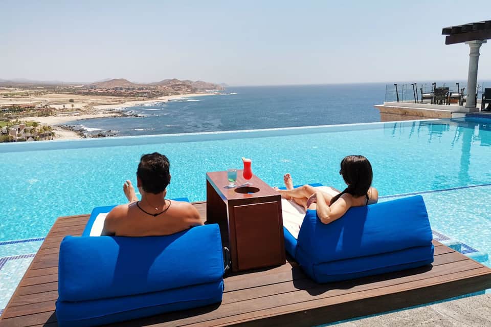 Two guests relaxing in blue lounge chairs by an infinity pool overlooking coastal cliffs and ocean