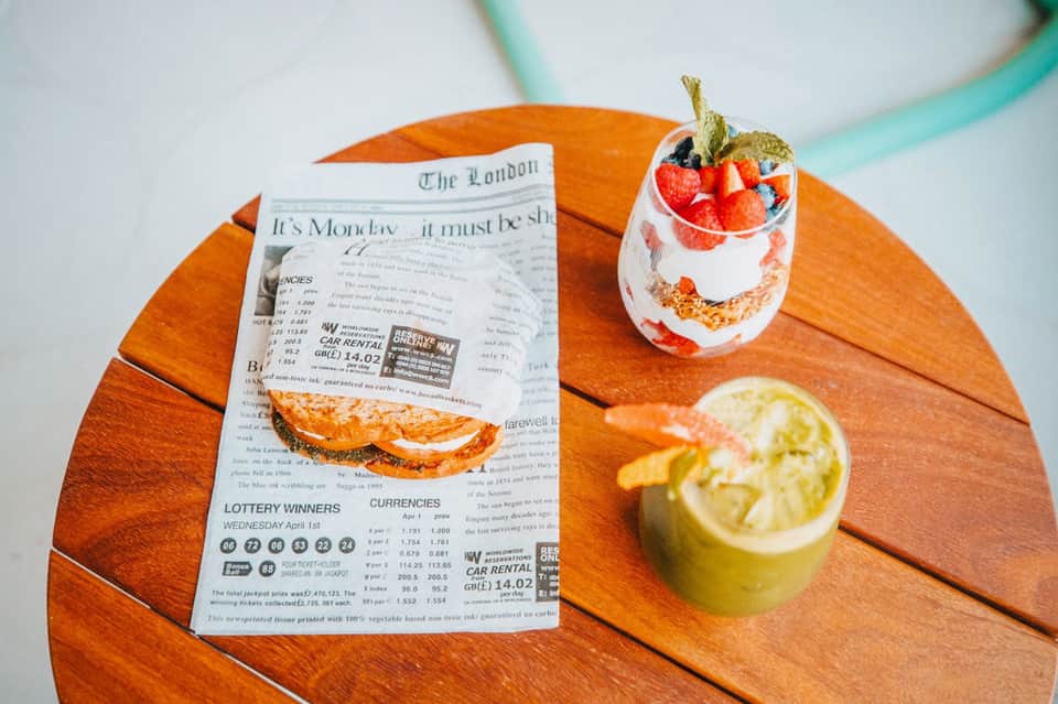 Wooden table with newspaper, layered dessert in glass, and lime slice garnish on turquoise background