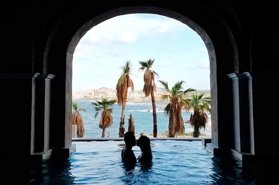 Couple in infinity pool framed by archway, palm trees, and turquoise ocean vista beyond