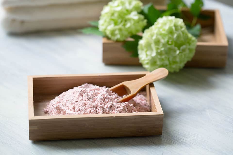 Pink clay powder in wooden bowl with wooden scoop and fresh green hops in background