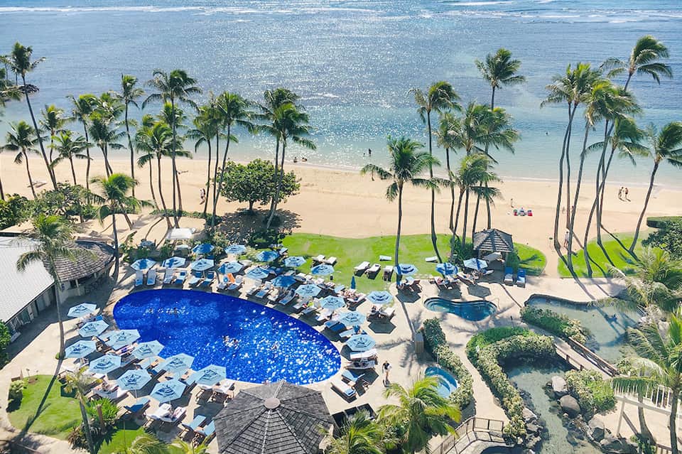 Aerial view of beachfront resort with kidney-shaped pool, loungers, and sandy beach with ocean