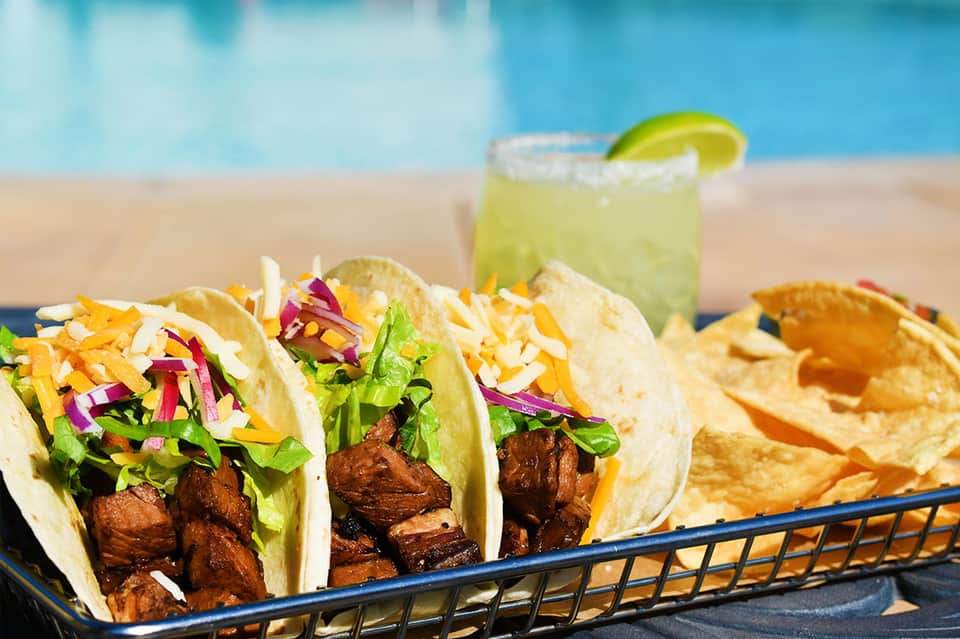 Three tacos with lettuce and purple cabbage by poolside with margarita and beach background