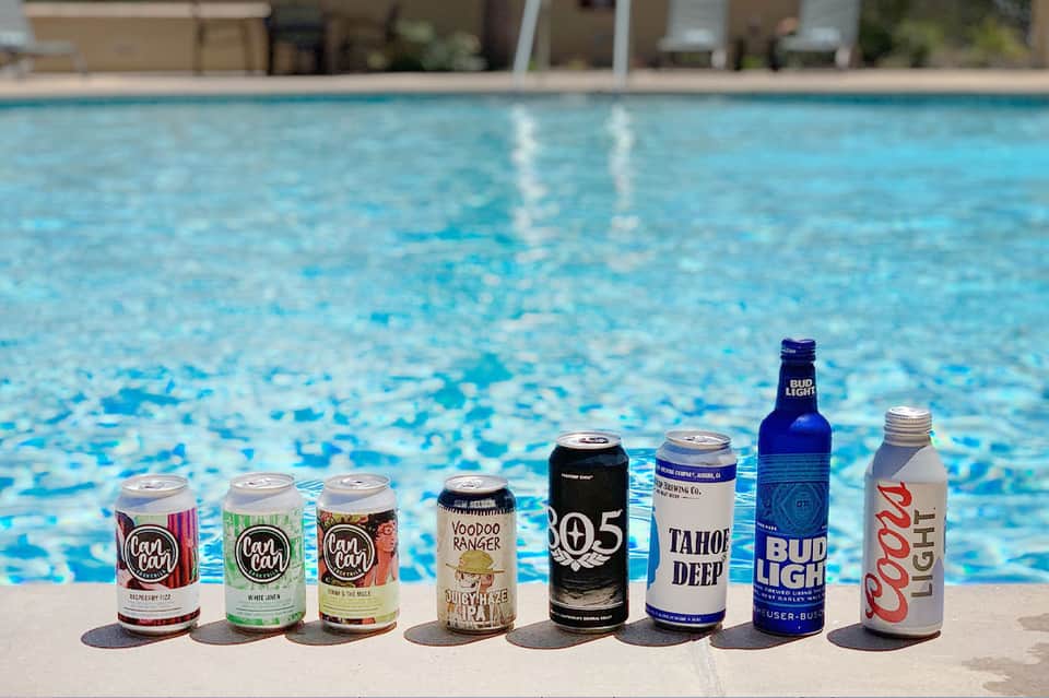 Eight beer cans and bottles lined up on pool deck edge overlooking a swimming pool