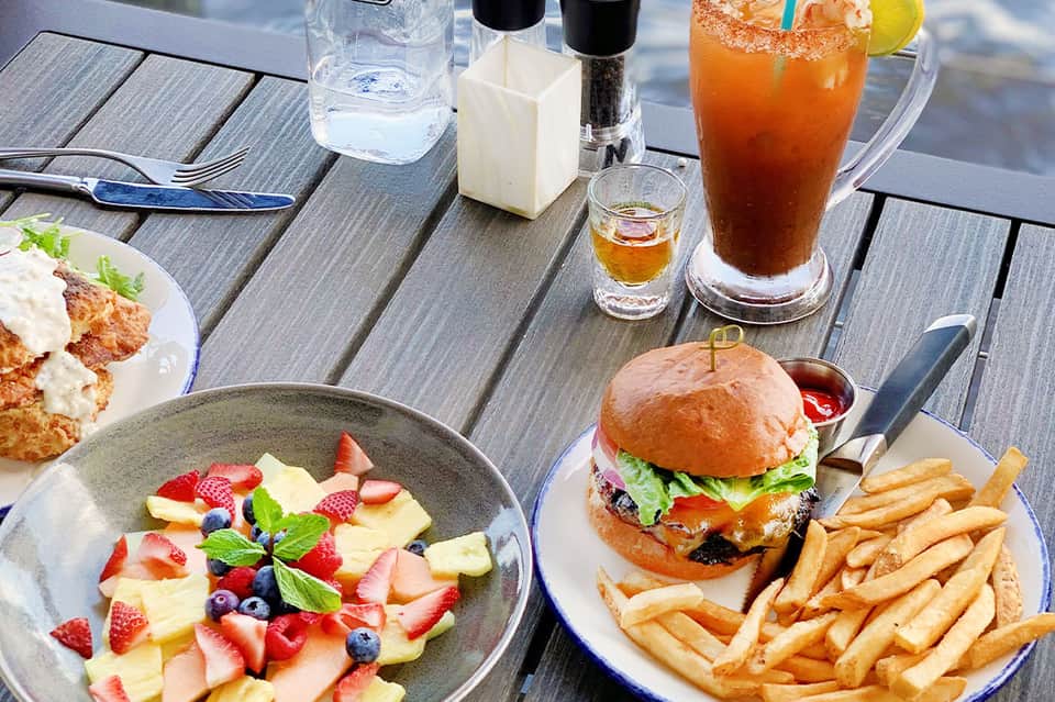 Outdoor dining table with fresh fruit salad, burger and fries, cocktails, and water on gray decking