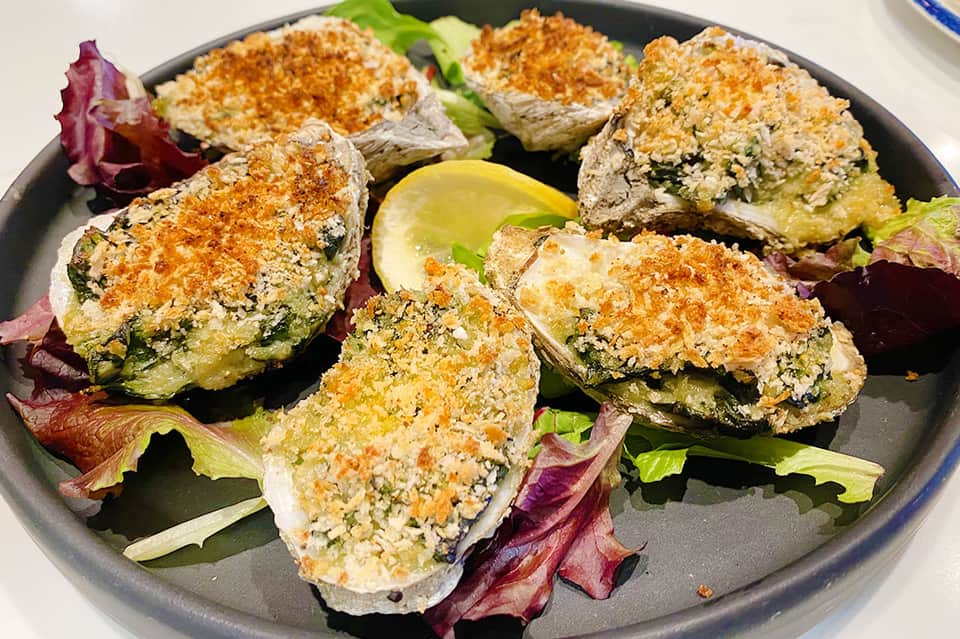Baked oysters with herb breadcrumb topping served on a dark plate with mixed greens and lemon wedge.