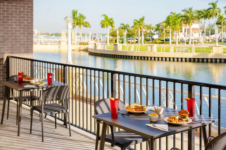 Waterfront dining terrace with tables overlooking a scenic river and palm trees in tropical setting