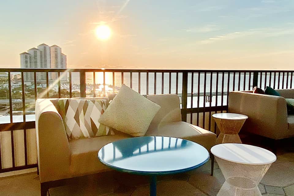 Beige lounge chair with pillows on a high-rise balcony overlooking sunset and distant city buildings