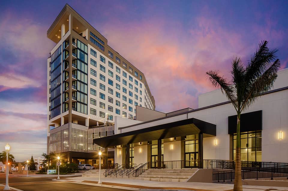 Modern hotel building at dusk with illuminated ground-floor entrance and palm trees
