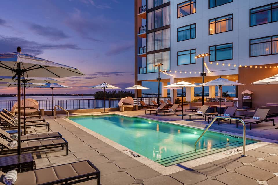 Illuminated rooftop pool at dusk with lounge chairs, umbrellas, and waterfront views