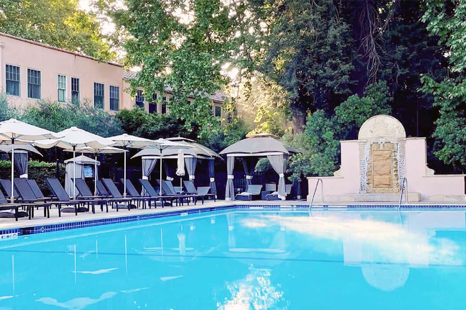Resort pool with lounge chairs, umbrellas, and cabanas surrounded by ivy-covered brick building and mature trees.