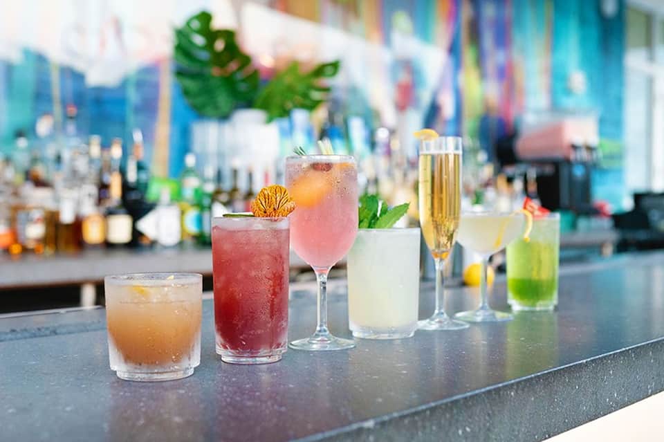 Colorful cocktails lined up on bar counter with tropical garnishes and blurred backdrop
