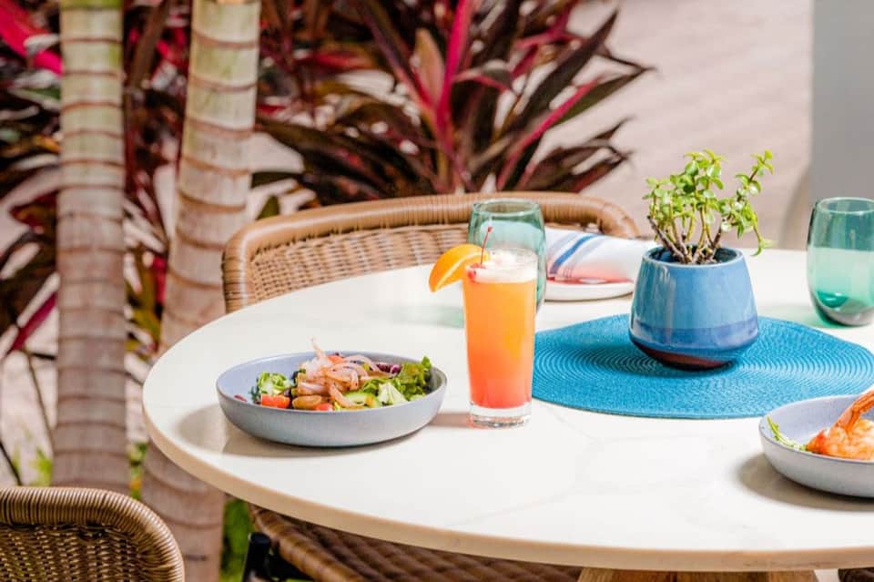 Outdoor dining table with salad, orange beverage, potted plant, and wicker chairs near tropical plants