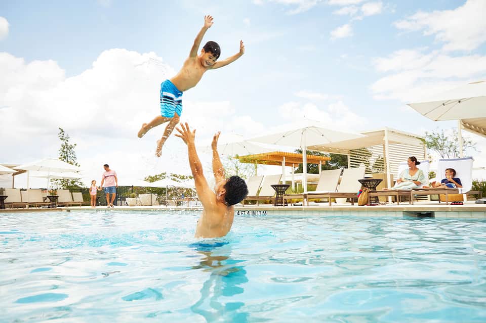 Father tosses young son in air while playing in resort swimming pool on sunny day with lounge chairs and umbrellas visible.