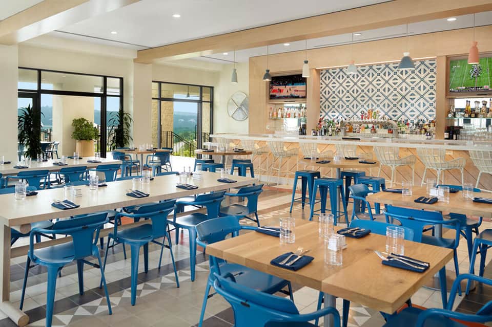 Modern hotel restaurant with blue chairs, wooden tables, natural light, and patterned backsplash behind the bar