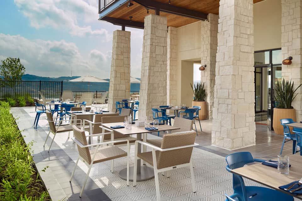 Upscale outdoor dining terrace with stone columns, wicker seating, blue chairs, and mountain lake views