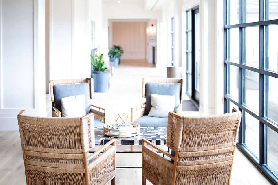 Modern lounge area with woven chairs, blue accents, and bright windows overlooking hallway