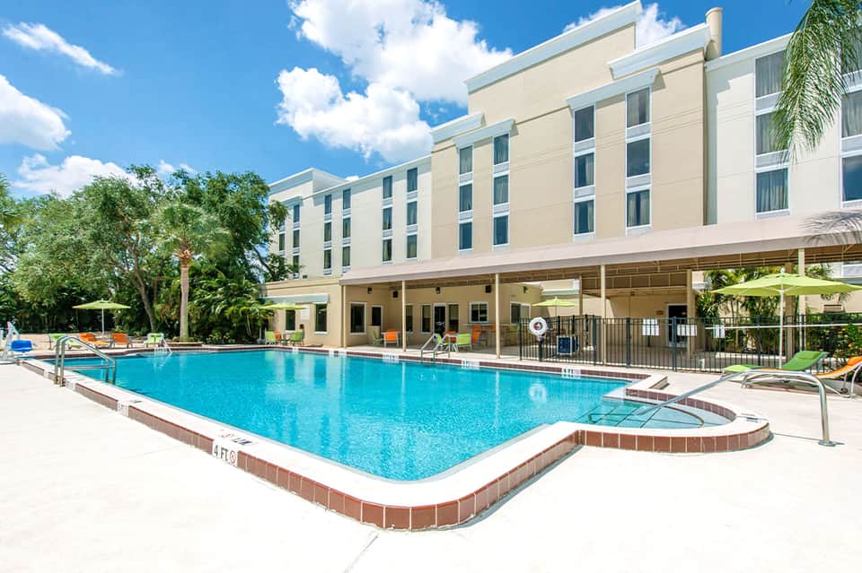 Large outdoor swimming pool with spa tub at hotel courtyard, surrounded by multi-story beige building and palm trees