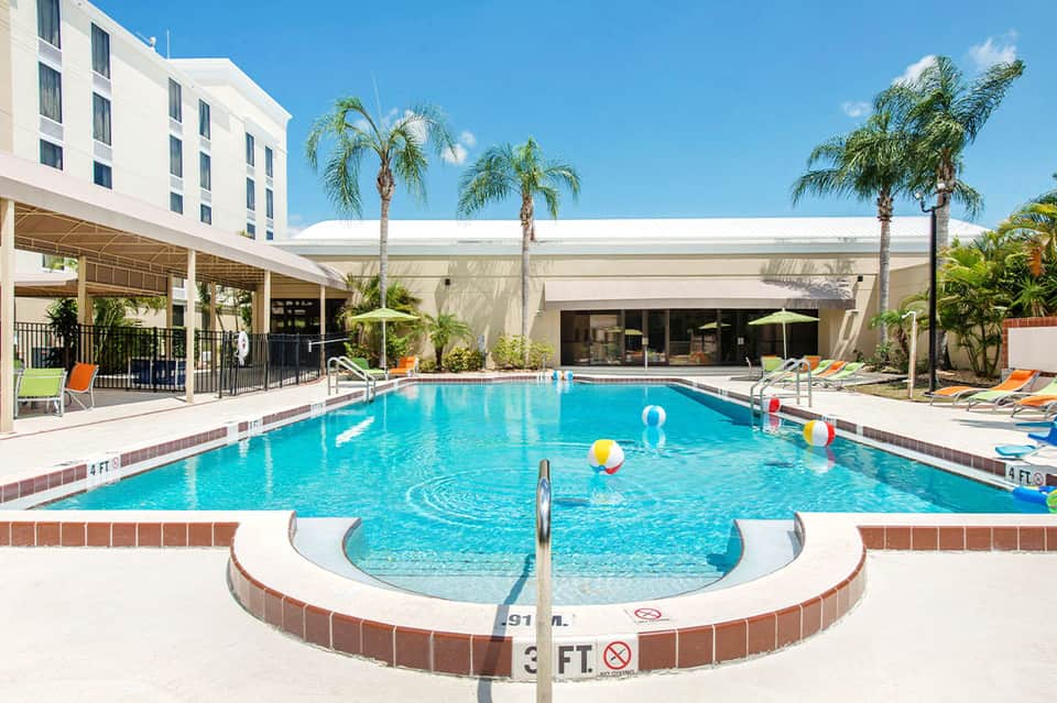 Resort outdoor pool with palm trees, loungers, white shade structure, and modern hotel building