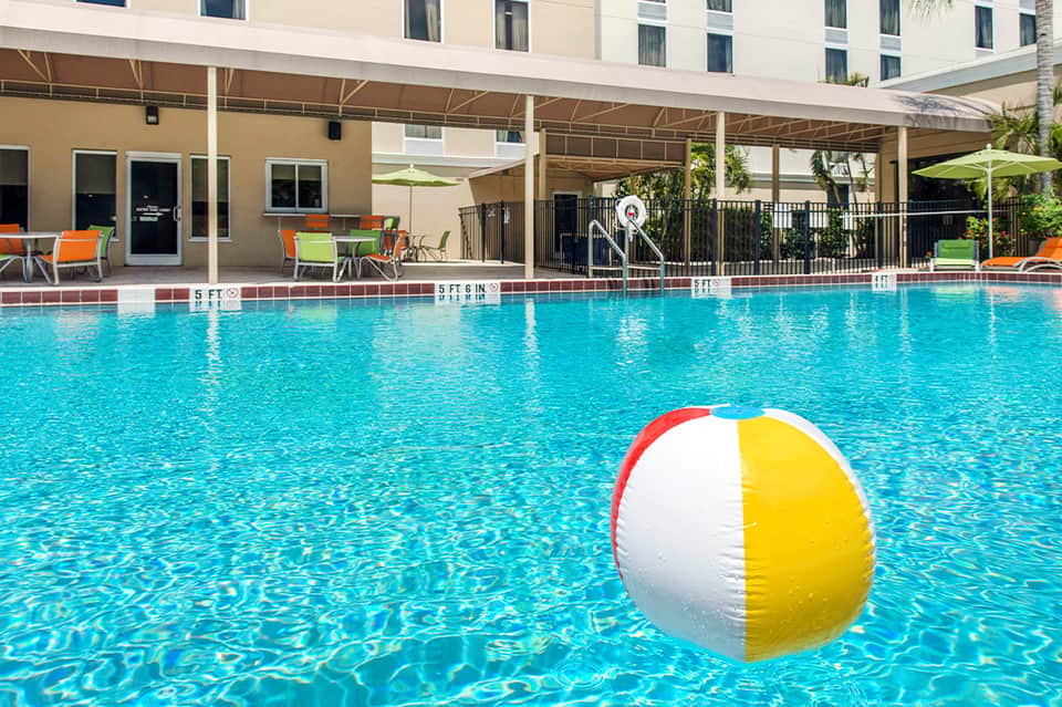 Resort pool with colorful beach ball, palm umbrellas, and lounging chairs under covered shade