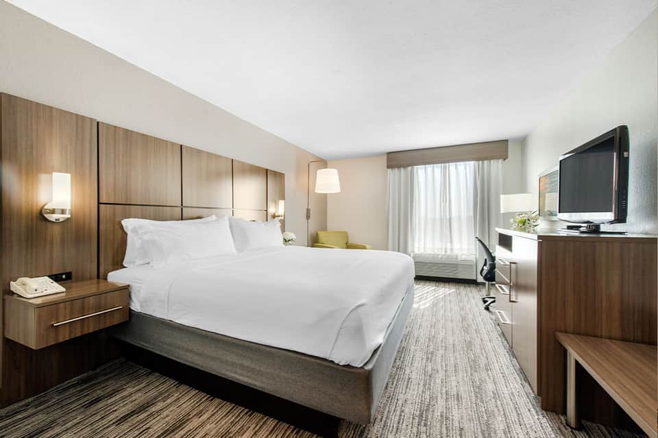 Modern hotel bedroom with wood headboard, white linens, TV, and natural light