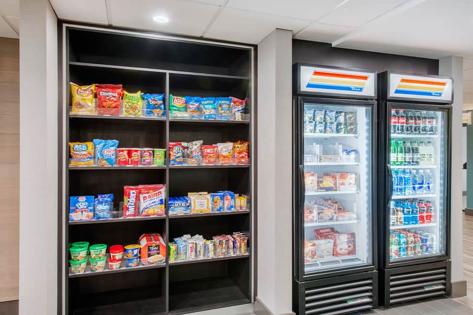Hotel snack bar with open shelving displaying chips and packaged goods, two refrigerated beverage coolers with drinks