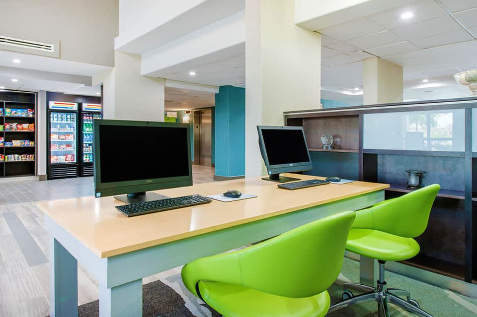 Modern hotel business center with lime green chairs, wooden desk, desktop computers, and refreshment area
