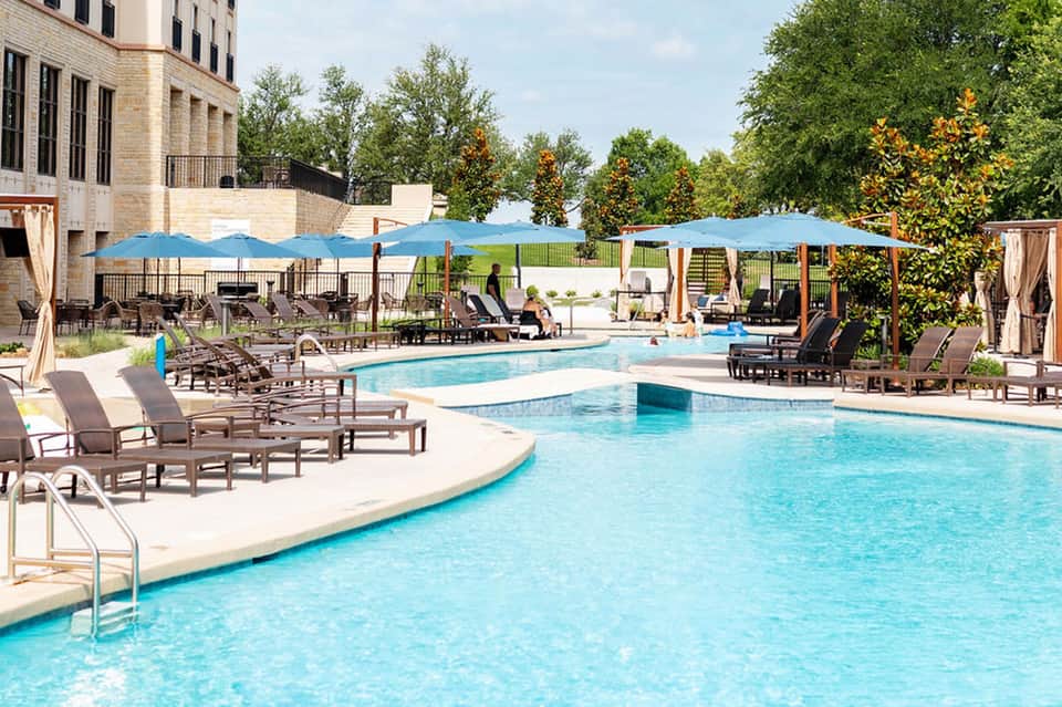 Resort pool area with blue umbrellas, lounge chairs, and manicured landscaping under clear sky