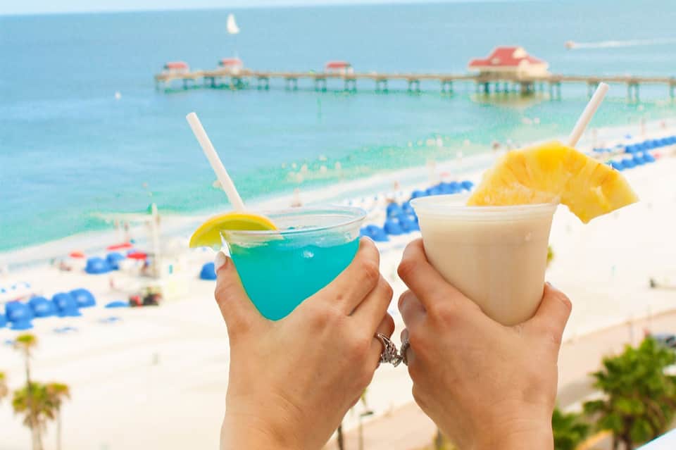 Two tropical cocktails with fruit garnish held up with beach and pier in background