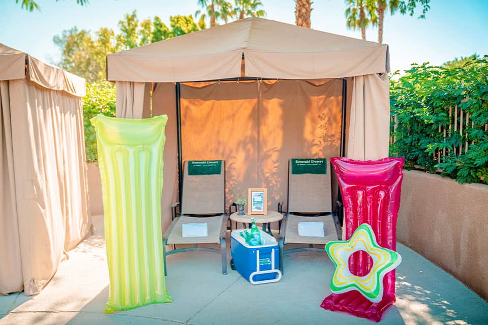 Outdoor cabana lounge area with tan canopy tent, lounge chairs, colorful pool floats, and tropical vegetation