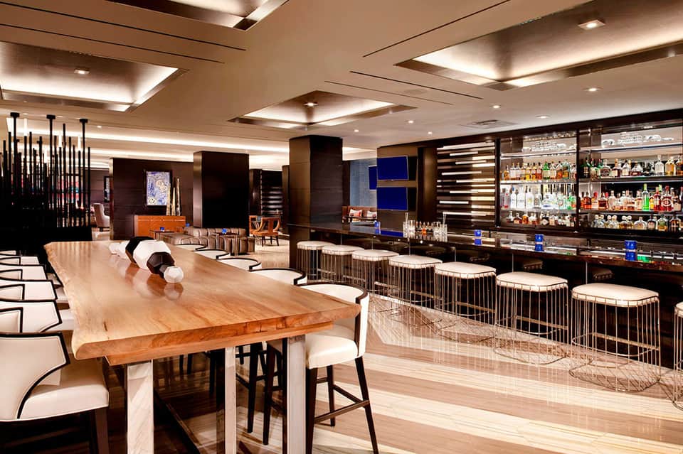 Upscale hotel bar lounge with wooden communal table, modern seating, and fully stocked liquor shelves
