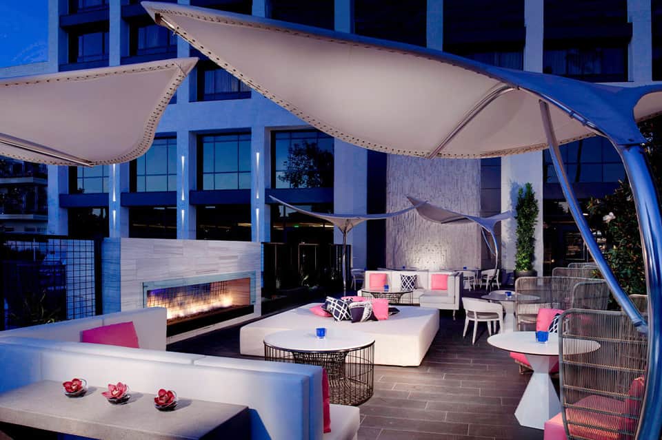 Modern rooftop lounge at dusk with fire pit, white seating, pink accents, and sailcloth canopies