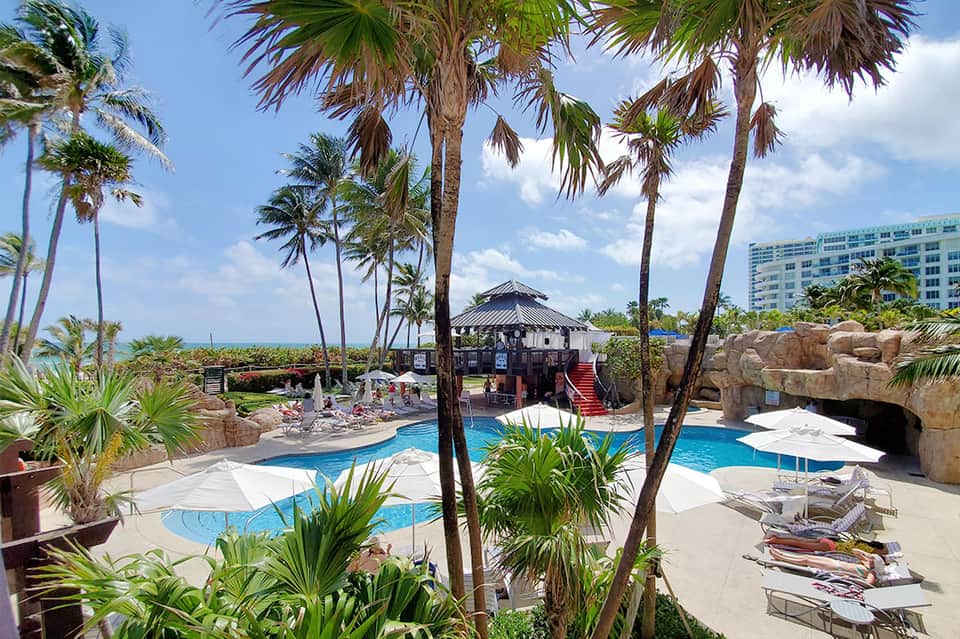 Resort pool area with palm trees, tiki bar, rock features, and tropical landscaping under blue sky