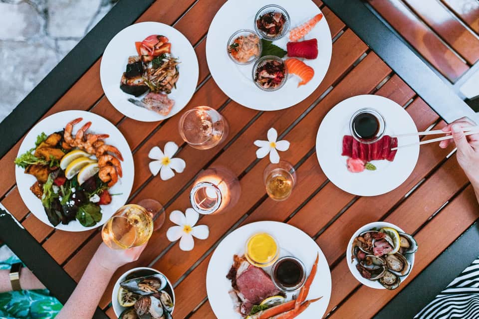 Overhead view of multiple white plates with seafood, meats, and appetizers on wooden deck with rosé wine and white flowers