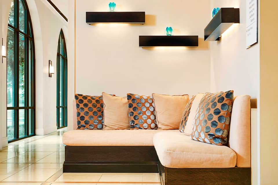 Modern living room with beige L-shaped sofa, patterned orange and blue pillows, black floating shelves with turquoise accents, and green-framed win...