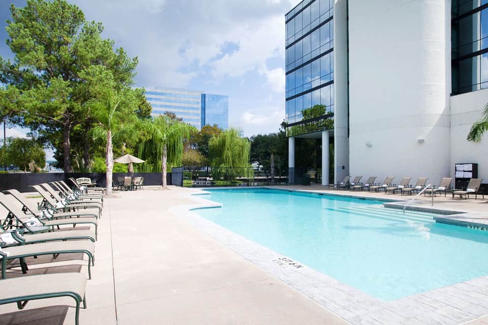 Contemporary hotel pool with clear turquoise water, sun loungers, mature trees, and modern white building facade.