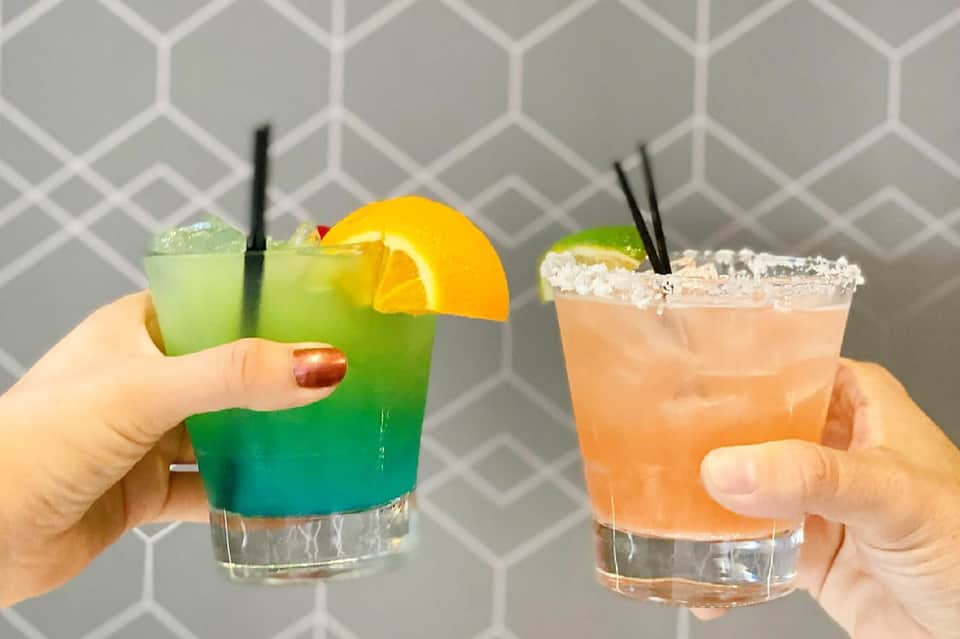 Two colorful cocktails with citrus garnishes and salt-rimmed glasses held by hands