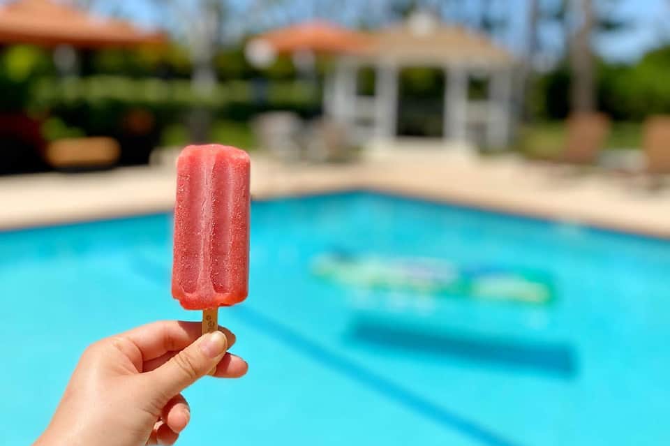 Hand holding red popsicle in front of bright blue resort swimming pool with loungers