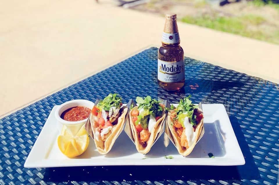 Four tacos with Modelo beer and salsa on a table with ocean view backdrop